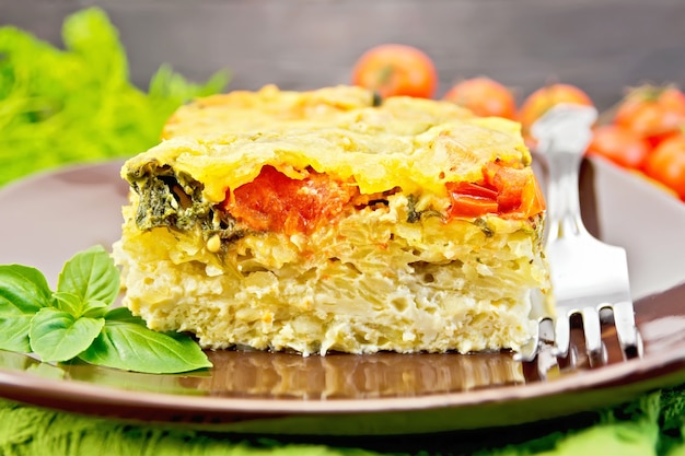 The pie of potatoes, cheese, tomato and spinach, filled egg with milk in a plate with a fork, a napkin on a dark wooden board