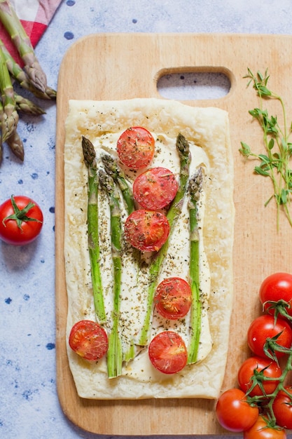 Pie with green asparagus cream cheese and tomatoes on a wooden cutting board on a light blue background