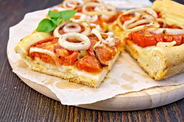 Pie with onions and tomatoes and basil on parchment and round board on a background of dark wooden board