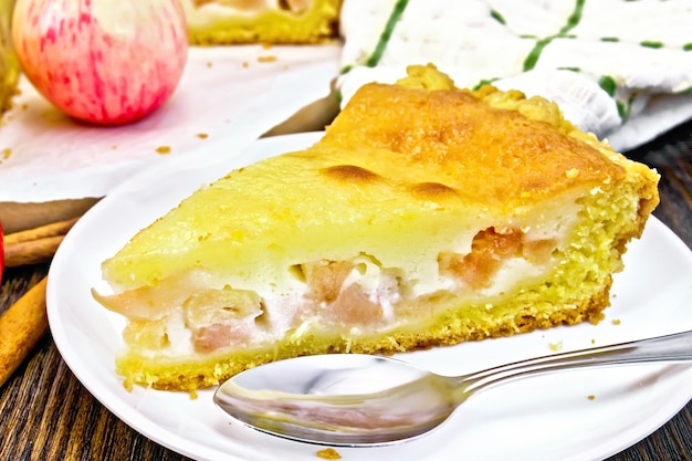 Piece of apple pie with sour cream sauce in a white plate, cinnamon, fruit and napkin on a background of a dark wooden board