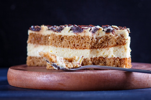 A piece of cake with milk and butter cream cutted with spoon on a wooden kitchen board, black table.