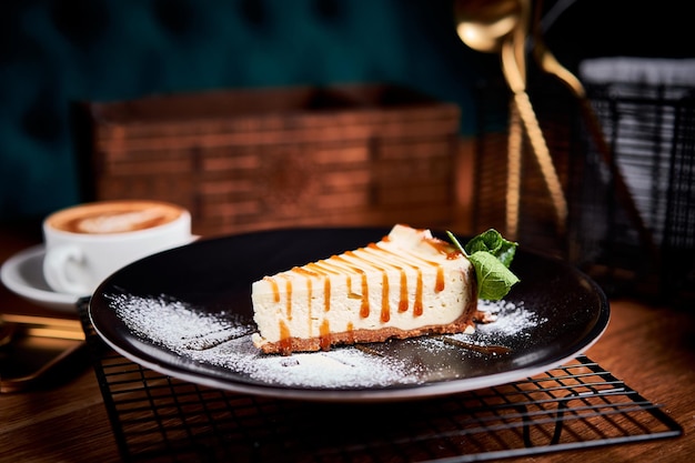 Piece of Cheesecake on black plate on a table with mint and a cup of coffee Side view