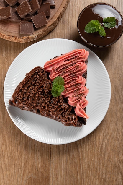 Piece of Chocolate Brownie Slice with Strawberry and Chocolate Icing Wooden table with mint and chocolate chips in the background Top view