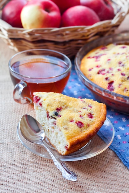 Piece of cowberry pie on glass plate and cup of tea and wicker basket with apples on background