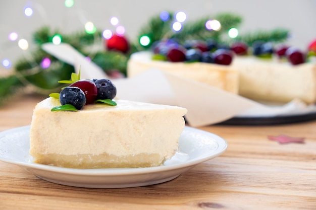 Piece of delicious cheesecake with cranberry and blueberry on plate with full cake in pine branches in bokeh lights on background Recipe of dessert for New Year and Merry Christmas festive treatment