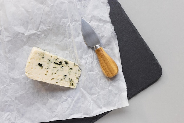 Piece of French blue roquefort cheese with blue mold and cheese knife on wrapping paper on black slate plate gray background Top view copy space