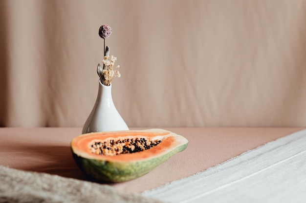 A piece of fresh papaya on it cut in half on a plate next to a vase with simple flowers Minimalism background natural concept