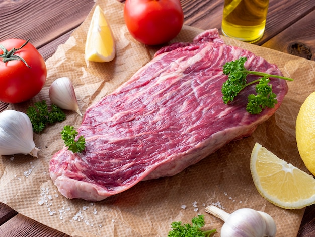 A piece of fresh raw beef lies on parchment surrounded by spices, herbs and vegetables. Side view. Food composition