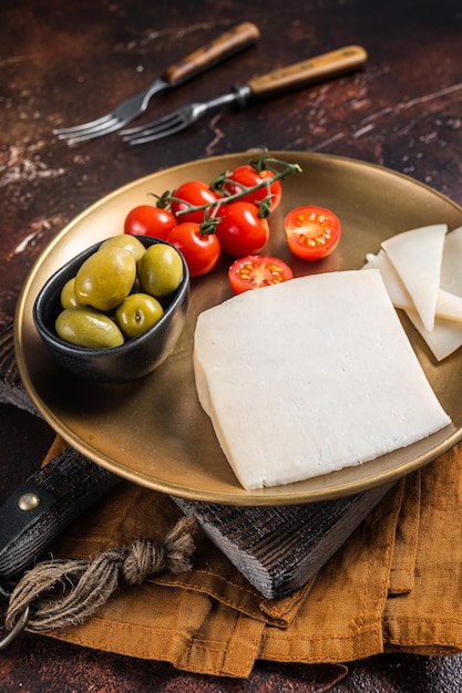 Piece of hard goat cheese in a plate with olives and tomato Dark background Top view