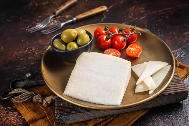 Piece of hard goat cheese in a plate with olives and tomato Dark background Top view