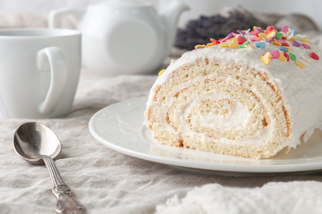 A piece of homemade biscuit roll with butter cream Decorated with colorful topping On a white plate In the background is a white teapot and a mug Closeup