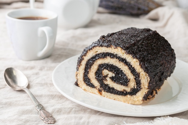 A piece of homemade biscuit roll with poppy seeds On a white plate In the background is a white teapot and a mug Closeup