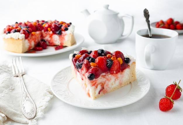 A piece of homemade cottage cheese berry pie Garnished with cream strawberries blueberries sea buckthorn berries On a white plate Next to the plate is a fork a napkin strawberries