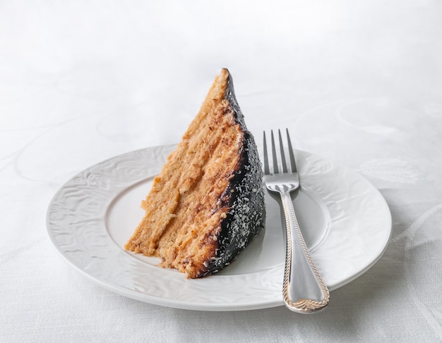 A piece of homemade multilayer biscuit cake Soaked in butter cream Garnished with chocolate icing and coconut On a white plate with a fork Closeup White background