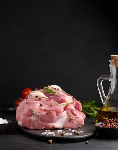 A piece of pork ham on a black board and spices olive oil salt rosemary branch and pepper