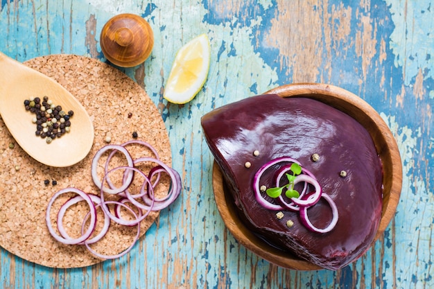 A piece of raw beef liver in a bowl, onion, lemon and spices for cooking on a wooden table. Top view