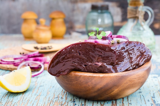 A piece of raw beef liver in a bowl and spices for cooking on a wooden table