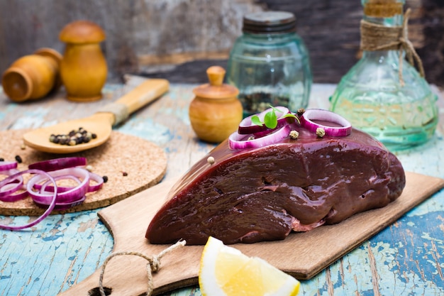 A piece of raw beef liver on a cutting board, onion, lemon and spices for cooking 