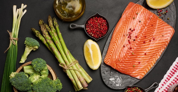 A piece of raw trout fillet on a wooden board next to asparagus and broccoli