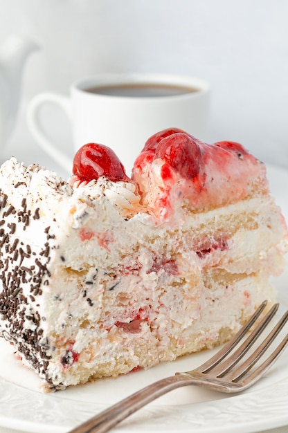 A piece of sponge cake with whipped cream and berries garnished with pieces of white chocolate Home made On a white plate In the background is a cake a teapot and a white mug