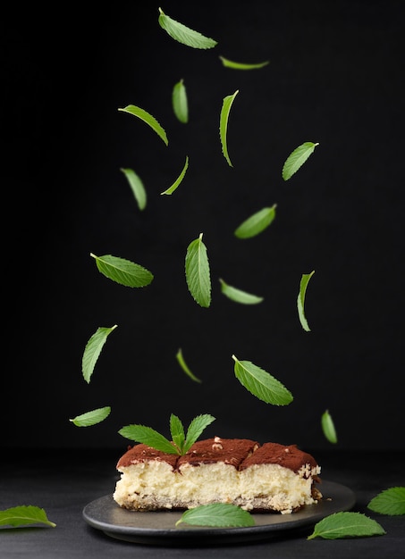 A piece of tiramisu sprinkled with cocoa flying green mint leaves over the dessert