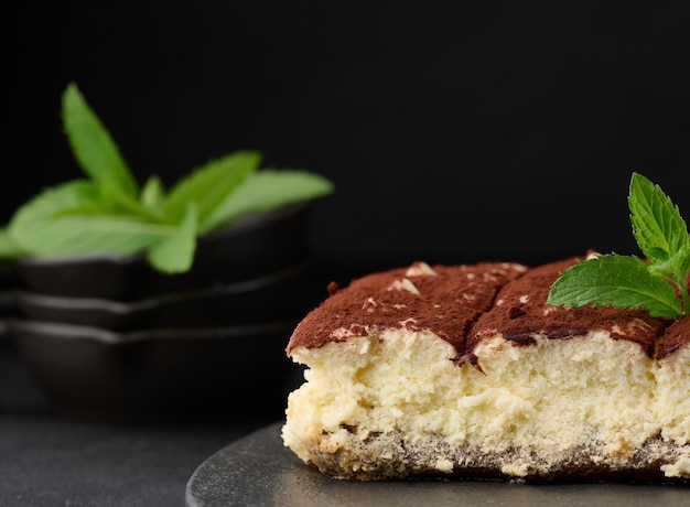 A piece of tiramisu sprinkled with cocoa on top of a sprig of fresh mint on a black background