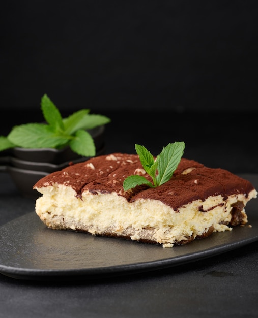 A piece of tiramisu sprinkled with cocoa on top of a sprig of fresh mint on a black background