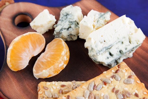 Pieces of blue cheese with mandarin and biscuits with seeds on a wooden board on a blue background Closeup of blue cheese