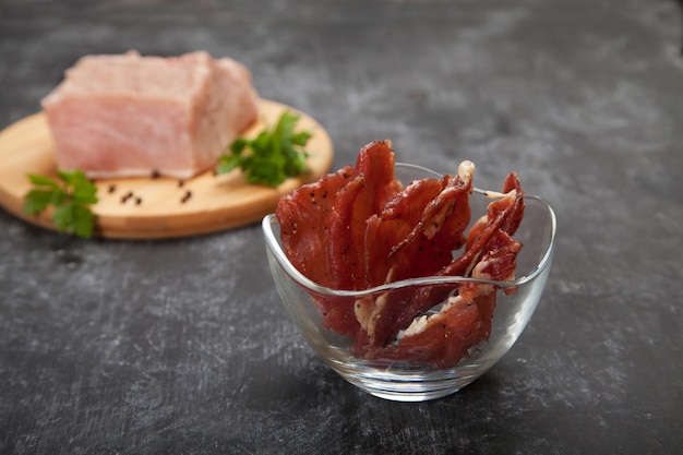 Pieces of jerky in a glass cup and raw meat on a cutting board on a black background.