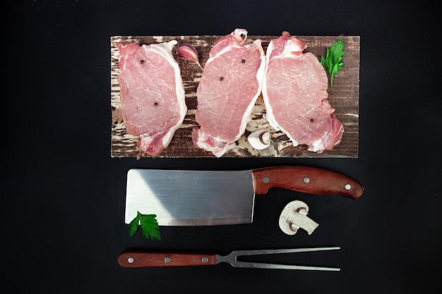 Pieces of raw roast beef meat for grilling on kitchen cutting board with parsley garlic and champignons knife and meat fork