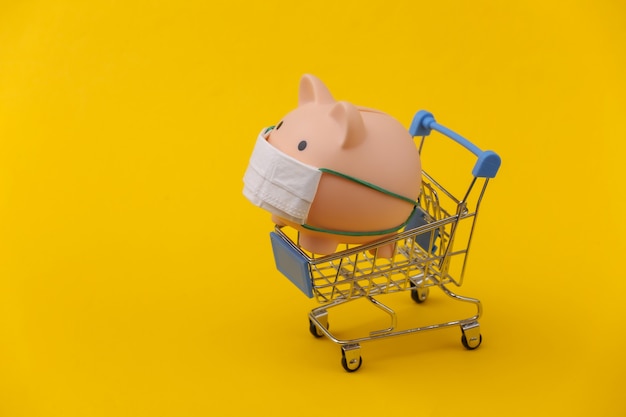 Piggy bank in a medical mask with a shopping trolley on yellow background. Shopping during covid-19, economic crisis, economy