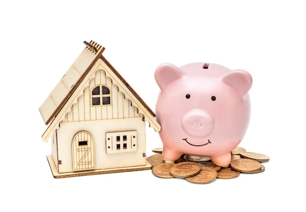 Piggy bank with model of house and heap of coins on white Savings money for buy house