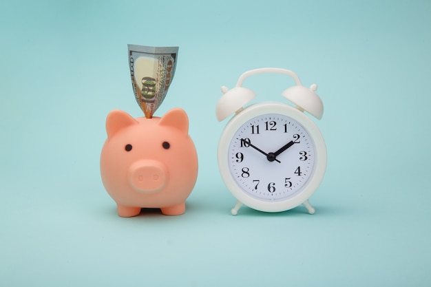 Piggy bank with money banknotes and alarm clock on blue background. Time is money concept