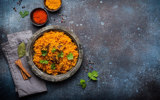 Pilaf - traditional Eastern dish made with rice, vegetables and meat served on vintage plate with fresh cilantro, sauces and spices on rustic background, top view and space for text