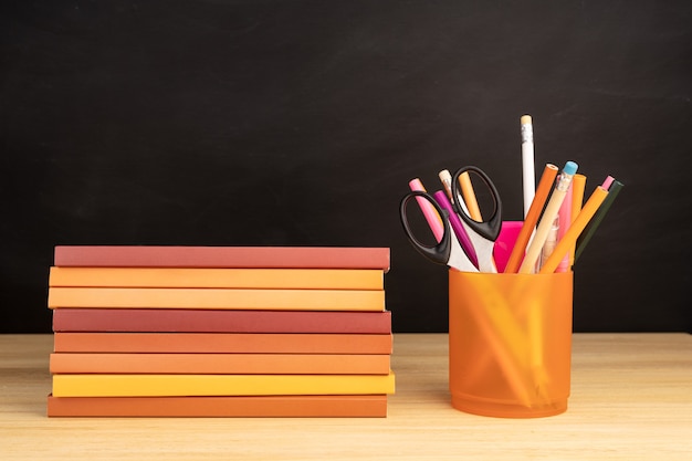 Pile of books and office supplies on wooden table and blackboard at background. Copy space