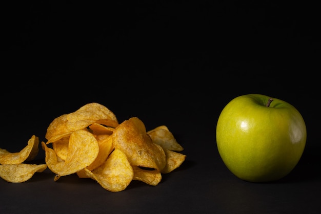 A pile of chips and a green apple lie side by side on a black background Healthy food concept