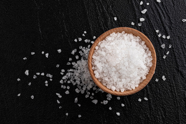 Pile of coarse sea salt in wooden bowl on black table, top view