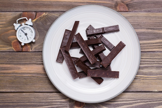 A pile of molded pieces of chocolate on a plate and an  alarm clock on a wooden table.  The concept of eating sweet time. Top view