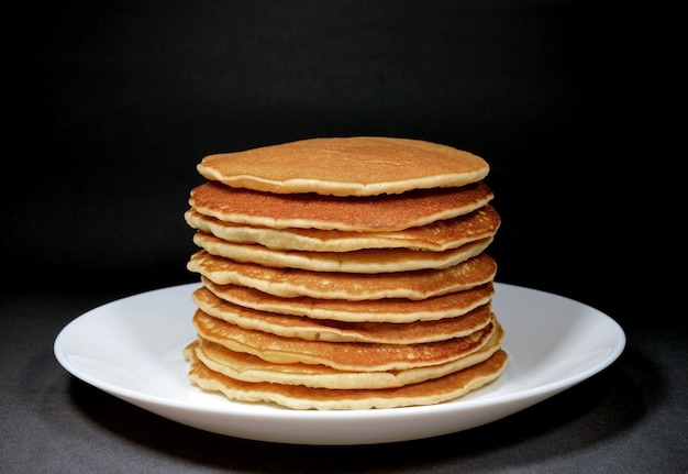 Piled up homemade plain pancakes served on white plate, isolated on black background