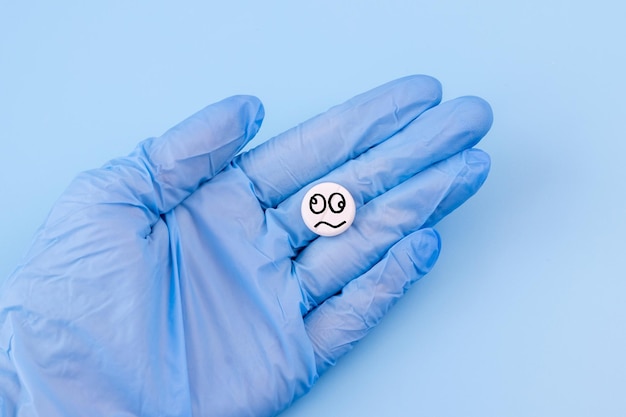 A pill with a crazy smiley in a hand in a medical glove on a blue background Drug concept