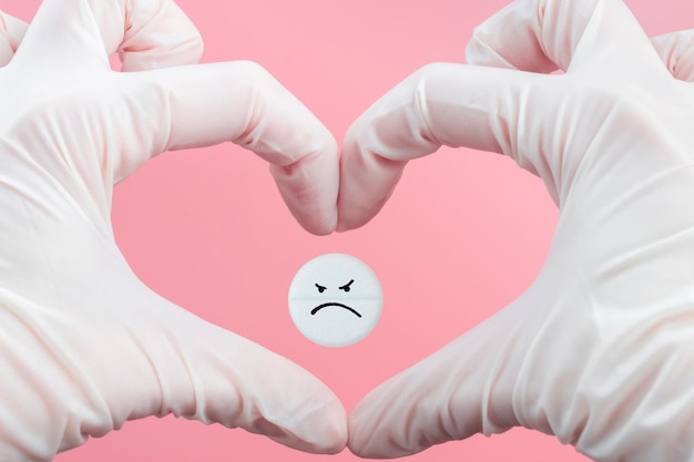 Pill with a sad emoticon in the heart from hands in medical gloves on a pink background Health care