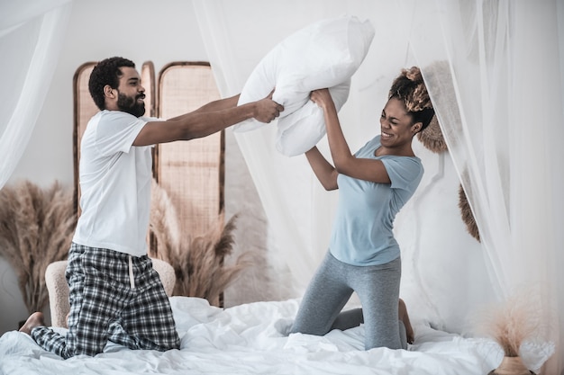 Pillow battle. Young adult black-skinned husband and wife in pajamas vigorously cheerfully fighting pillows on bed in bedroom