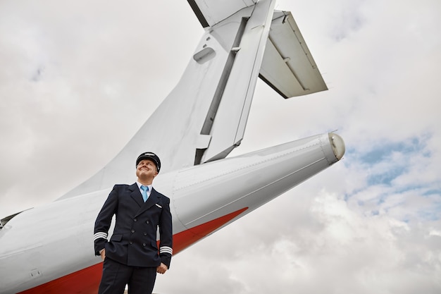 Pilot standing on runway on airplane jet background. Modern passenger plane. Bottom view of thoughtful european man with hands in pockets wear uniform. Civil commercial aviation. Air travel concept