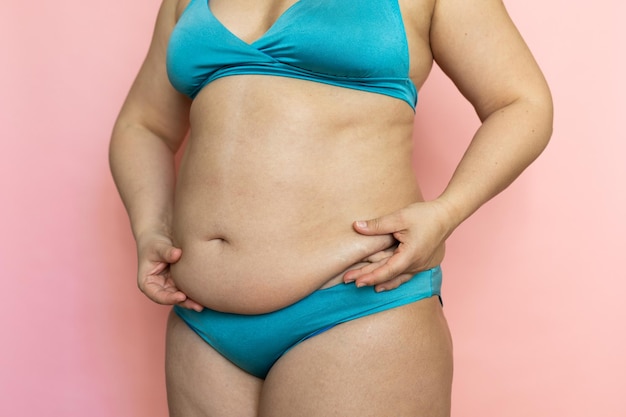 Pinch woman sagging belly with fingers closeup folds on stomach loose skin and cellulite Naked overweight plus size girl on pink background in blue underwear Concept of dieting and body control