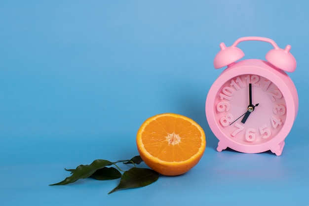 Pink alarm clock and half an orange on a blue background. The concept of summer drinks, healthy eating. Copy space.