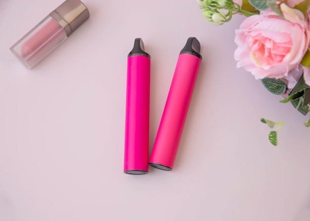 On a pink background there are two pink electronic cigarettes next to lipstick and pink flowers top view