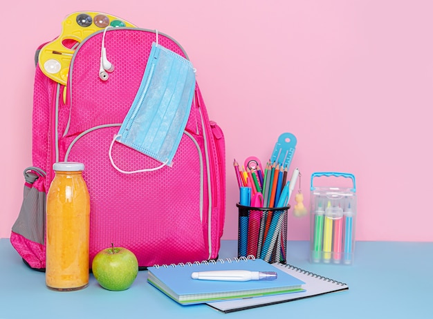 Pink backpack with medical mask, school supplies and lunch of fruit smoothie and apple