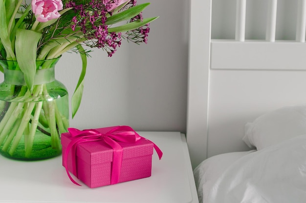 Pink box present and vase with spring flowers on bedtable near bed Scandinavian Interior