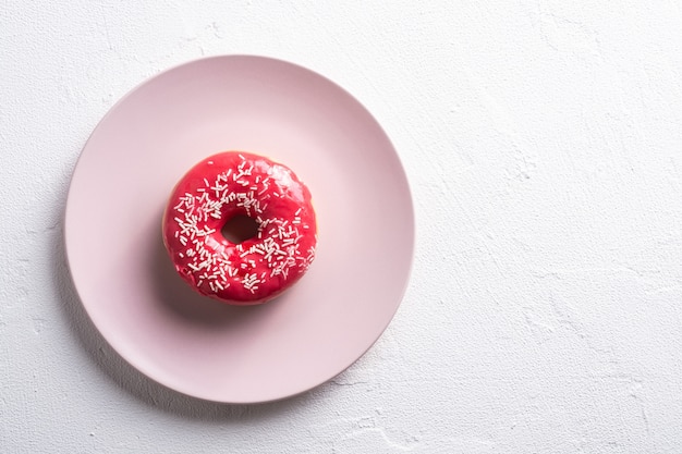 Pink donut with sprinkles on pink plate, sweet glazed dessert food on white concrete textured table, top view copy space