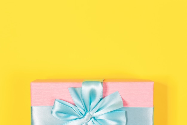 Pink gift box tied with blue ribbon with bow at the top on yellow background. Copy space for text. Minimal flat lay.